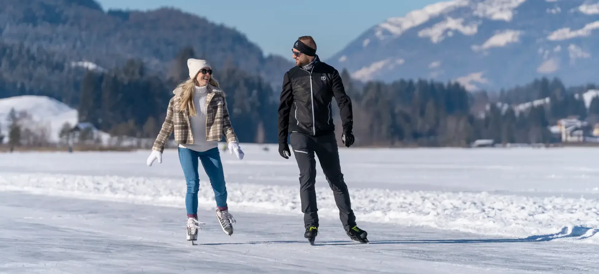 Paar beim Eislaufen auf einem zugefrorenen See mit schneebedeckten Bergen im Hintergrund, Winterfreizeitaktivitäten im Freien.