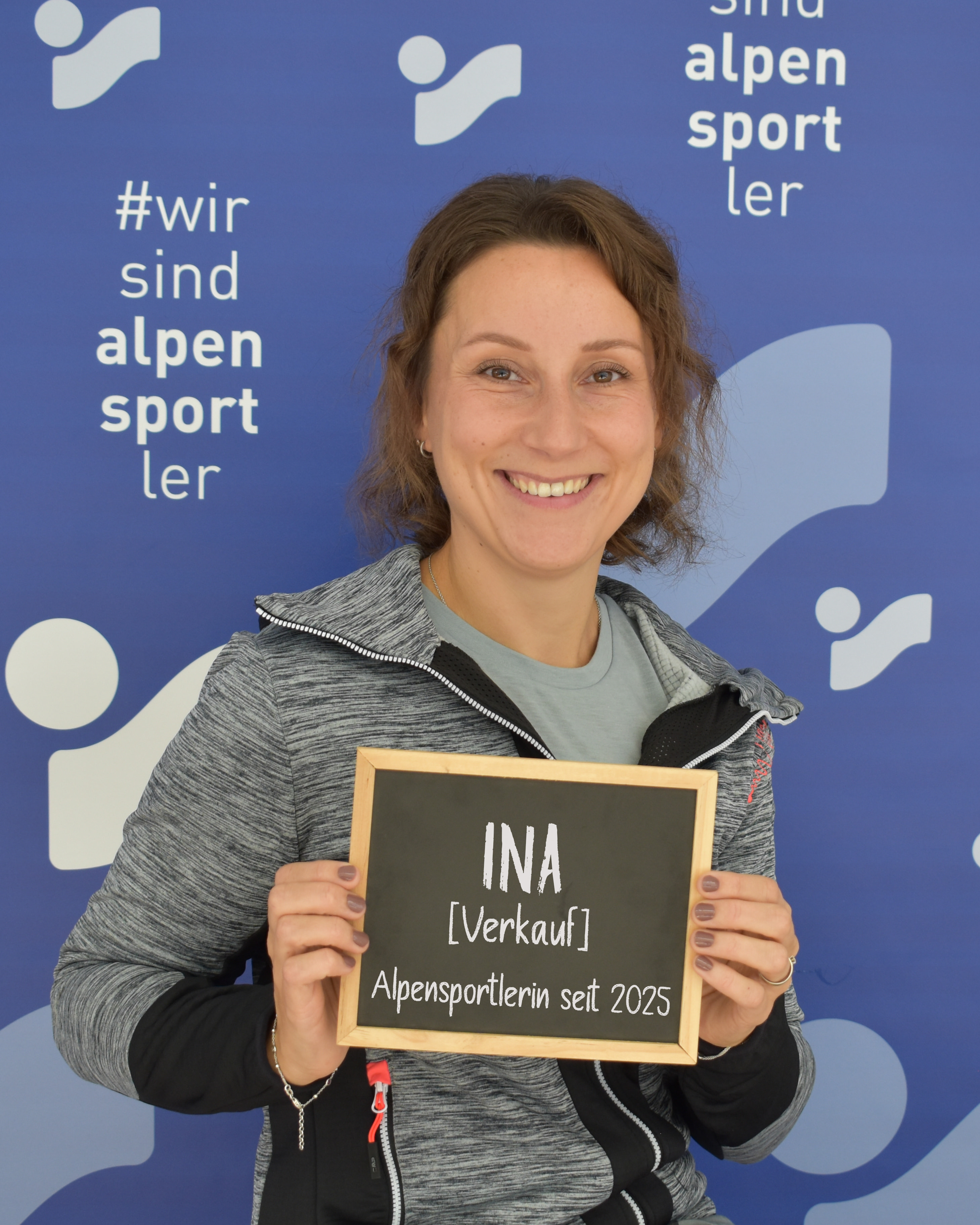 Lächelnde Frau hält ein Alpensportlerin-Schild mit dem Text "INA Verkauf".