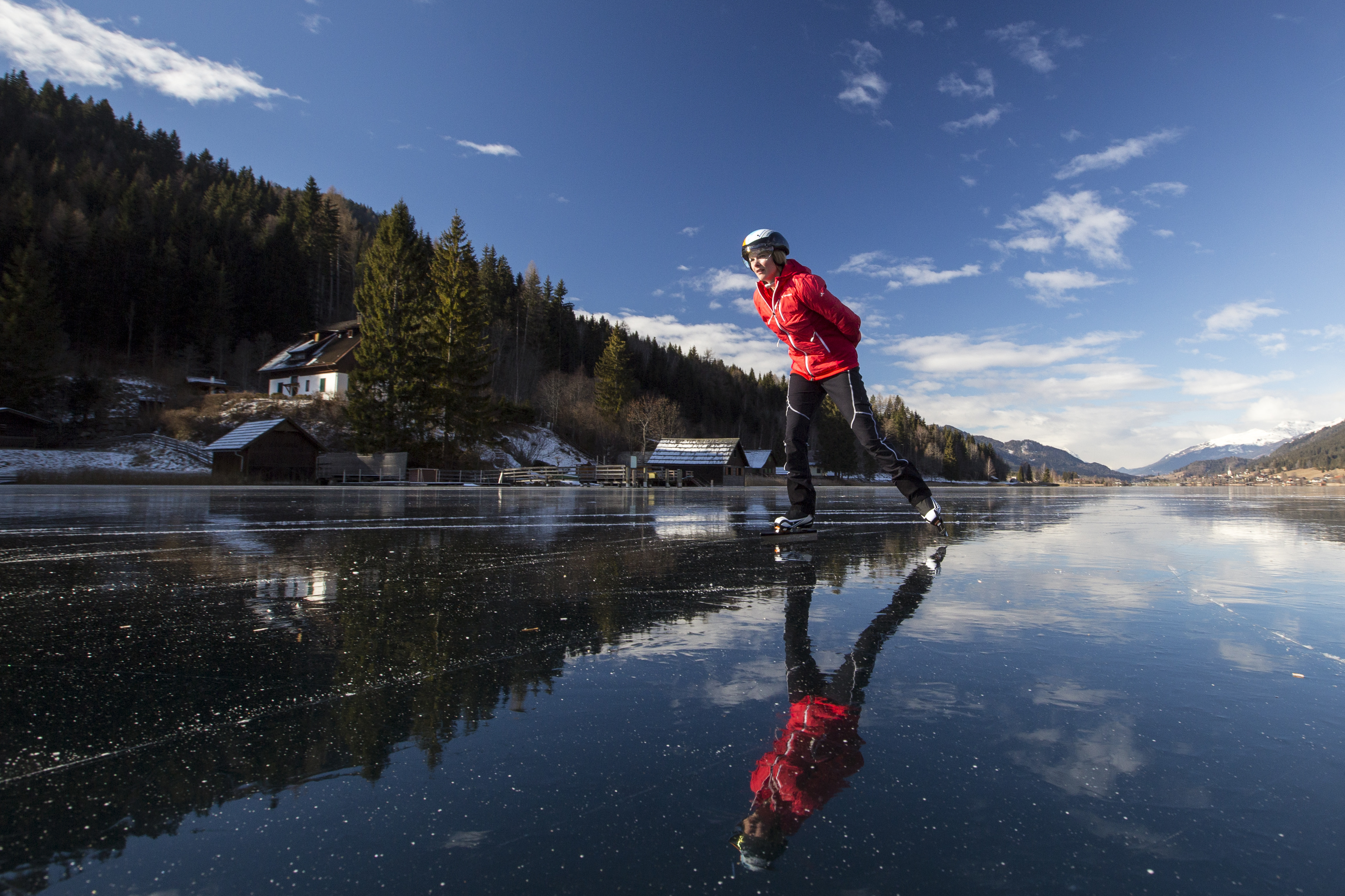 Frau beim Eislaufen auf einem zugefrorenen See vor dem Hintergrund von Bergwäldern und blauem Himmel. Wintersport.