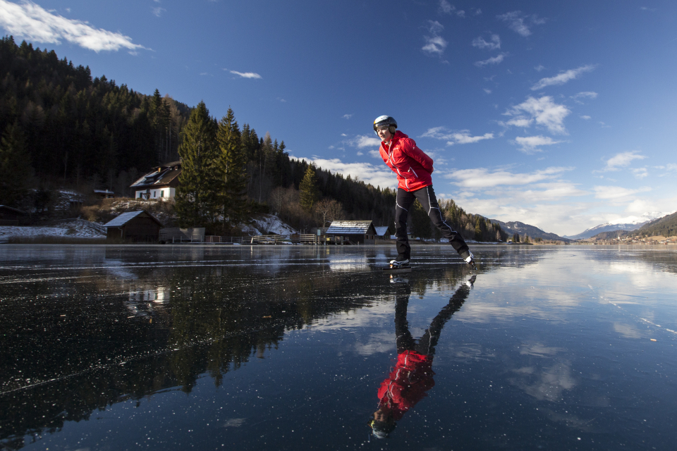 Frau beim Eislaufen auf einem zugefrorenen See vor dem Hintergrund von Bergwäldern und blauem Himmel. Wintersport.