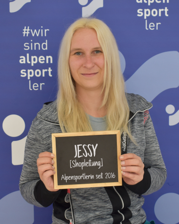 Blonde Frau hält ein Schild: Jessy, Filialleiterin bei Alpensportl seit 2016. Mitarbeiterprofil.