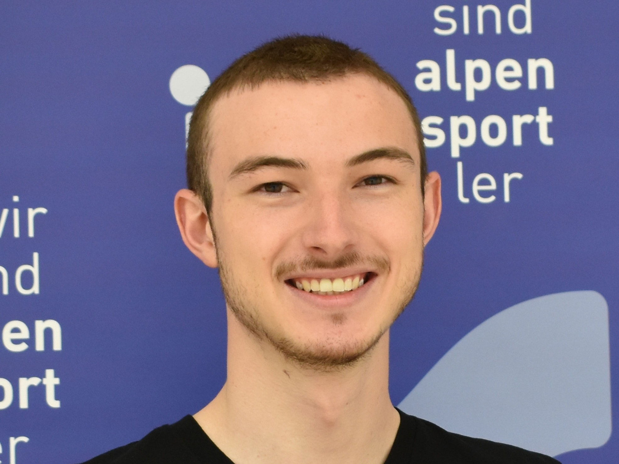 Lächelnder junger Mann (Porträt); Alpen Sport Ler Logo im Hintergrund.