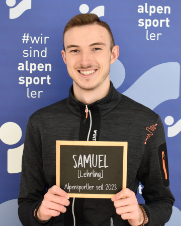 Lächelnder junger Mann, Samuel, hält ein Schild für das Alpensportl 2023 Ausbildungsprogramm.
