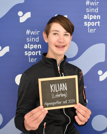 Der junge Alpensportler Kilian hält ein Schild mit der Aufschrift: "Kilian [Lehrling] Alpensportler seit 2024" vor einem gebrandeten Hintergrund.