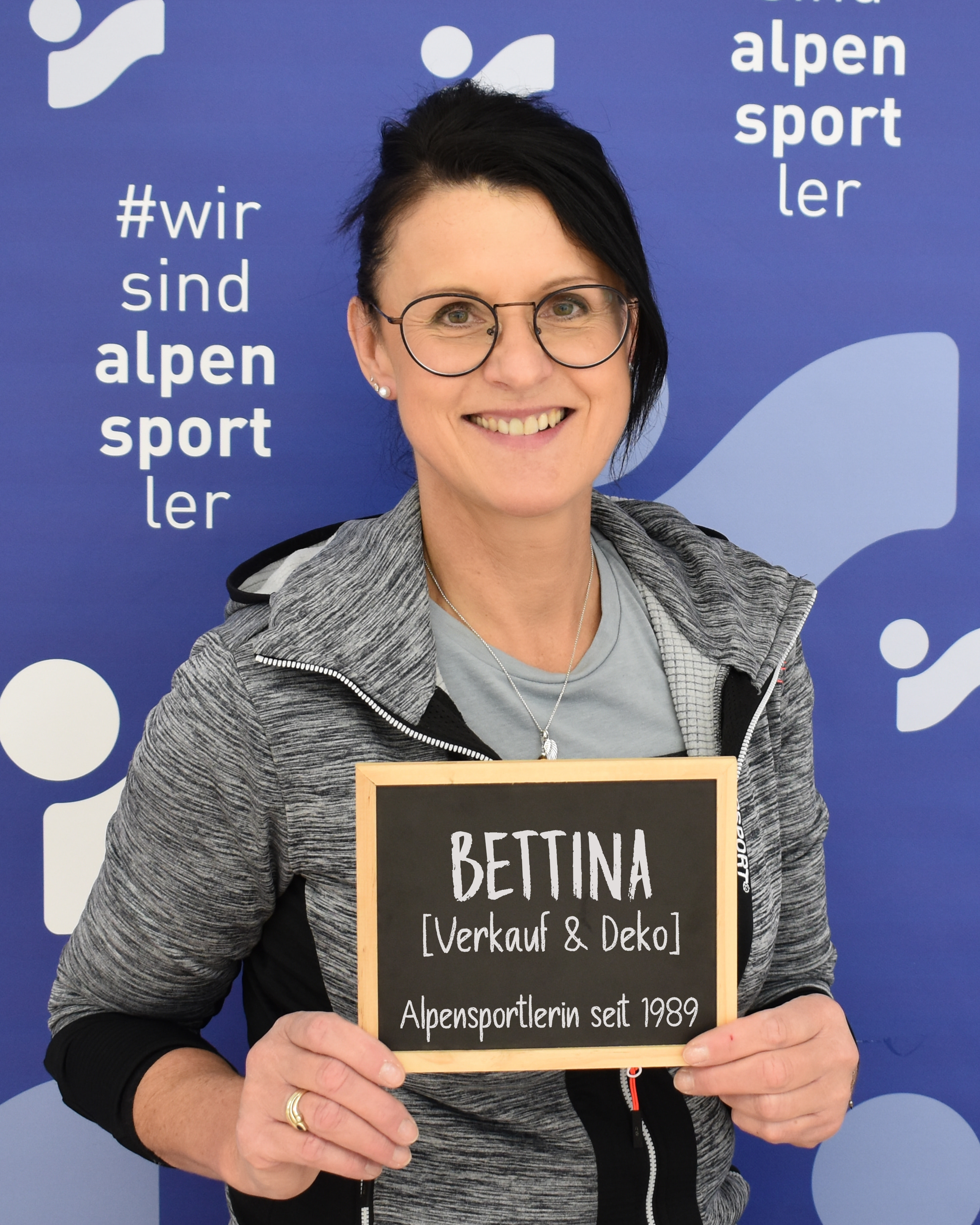 Lächelnde Frau hält ein Bettina-Namensschild für ein Mitglied des Verkaufs- und Dekorationsteams des Alpensportler-Skishops.