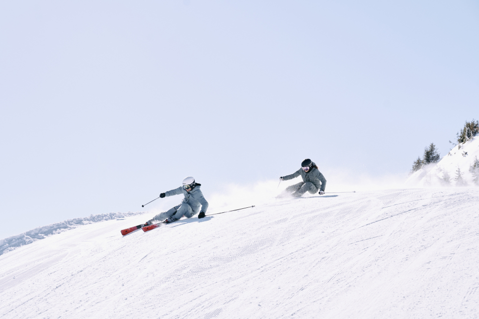 Zwei Personen fahren an einem sonnigen Tag Ski bergab und genießen Wintersport und Erholung in den Bergen.