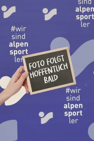 Hand, die ein Schild mit der Aufschrift "Foto folgt hoffentlich bald" vor blauem Alpensport-Hintergrund hält.