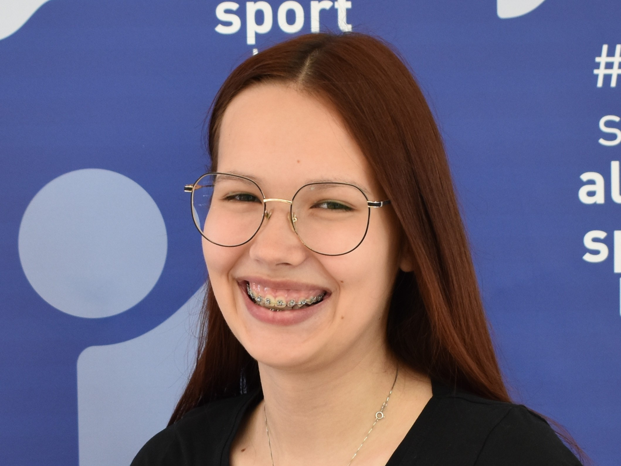 Lächelndes Teenager-Mädchen mit Zahnspange und Brille vor einem blauen Sport-Hintergrund.