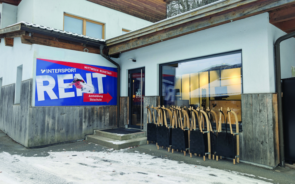 Außenansicht eines Skiverleihgeschäfts mit Schlitten, Intersport-Schild und Comic-Pinguin in verschneiter Winterlandschaft.