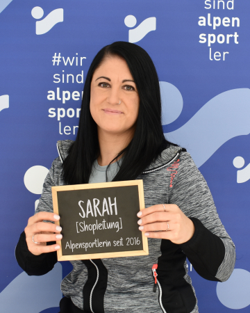 Sarah, Mitarbeiterin von Alpensport, hält ein Schild mit ihrem Namen und ihrer Position.