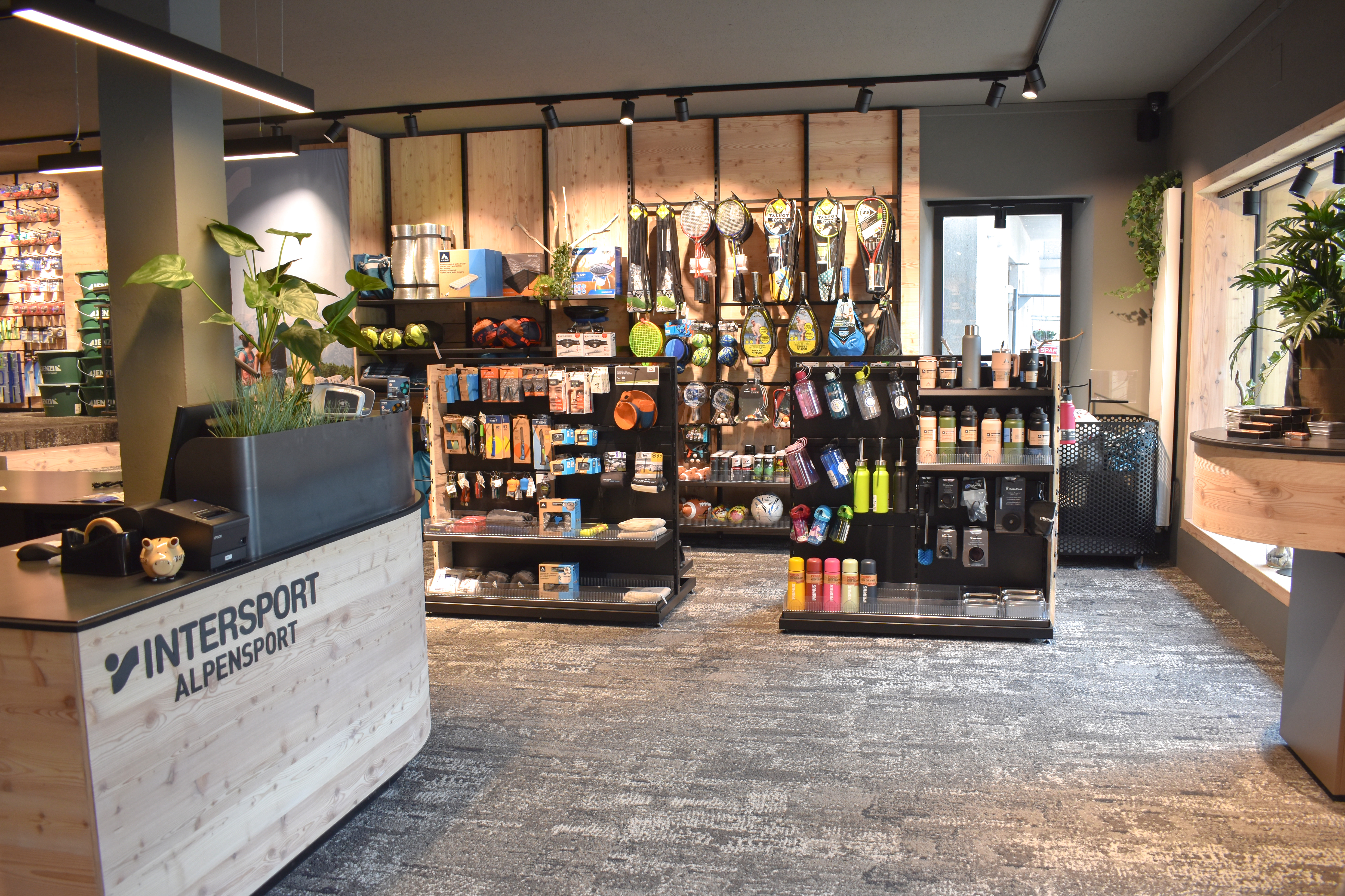 Foto der Camping-Abteilung bei INTERSPORT Alpensport Weissensee 15er Haus mit Trinkflaschen, Campinggeschirr, Campingstühle, Schlafsäcke, Unterlagsmatten, sowie der Sport-und Spiel Abteilung mit Federball, Tischtennis, Volleyball und verschiedenen Strandspielen.