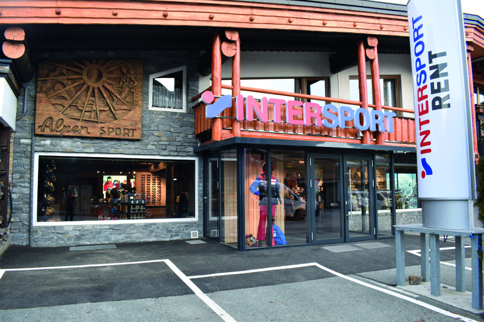 Außenansicht des Intersport Rent Skigeschäfts mit Almen Sport-Schild, Skiausrüstung im Inneren sichtbar.