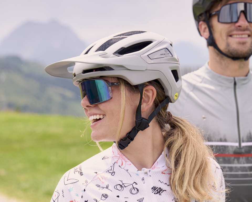 Frau mit grauem Mountainbike-Helm mit Visier, lächelnd im Freien mit Radfahrer im Hintergrund.