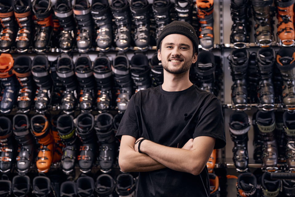 Lächelnder Mann steht in einem Skigeschäft vor einer Wand mit Skischuhen zum Verkauf oder zur Miete.