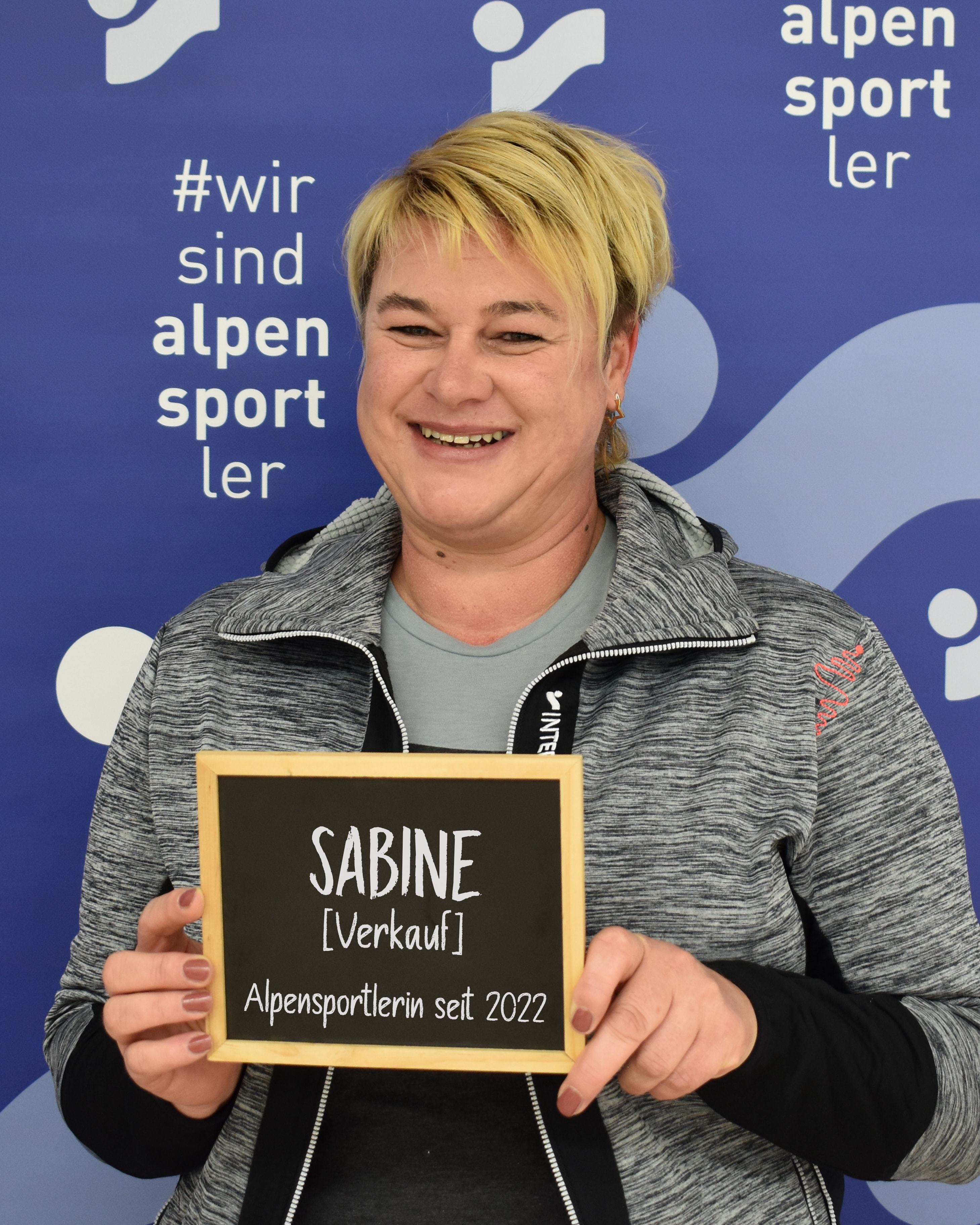 Lächelnde Frau hält ein "Sabine Verkauf" Schild für Alpensport.