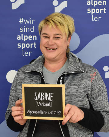 Lächelnde Frau hält ein "Sabine Verkauf" Schild für Alpensport.